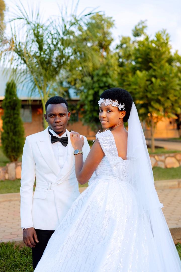 Bride Donatha MUKESHIMANA in a white wedding dress and groom Elie NSENGIMANA in a white suit and black pants, smiling together on their wedding day at ADEPER Church Nyamata.