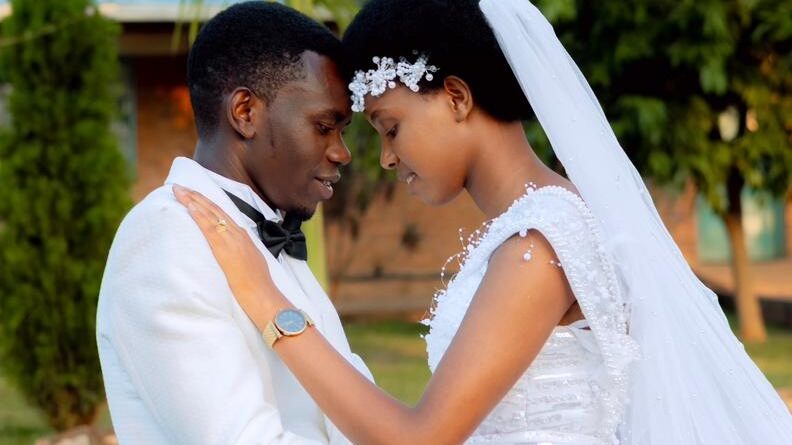 Bride Donatha MUKESHIMANA in a white wedding dress and groom Elie NSENGIMANA in a white suit and black pants, smiling together on their wedding day at ADEPER Church Nyamata.